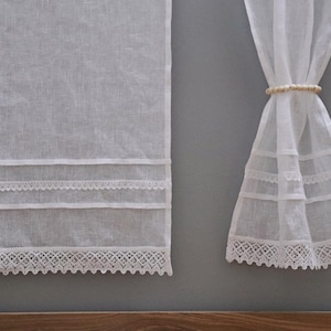 Peut inclure: Rideaux en lin blanc avec garniture en dentelle d&eacute;corative. Les rideaux sont suspendus contre un mur gris. La garniture en dentelle se trouve en bas et en bandes horizontales sur les panneaux de rideaux. Un embrasse-rideau perl&eacute; maintient un panneau.