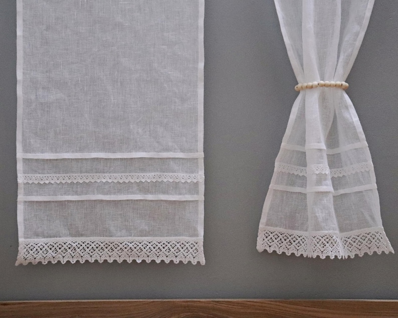 Peut inclure: Rideaux en lin blanc avec garniture en dentelle d&eacute;corative. Les rideaux sont suspendus contre un mur gris. La garniture en dentelle se trouve en bas et en bandes horizontales sur les panneaux de rideaux. Un embrasse-rideau perl&eacute; maintient un panneau.