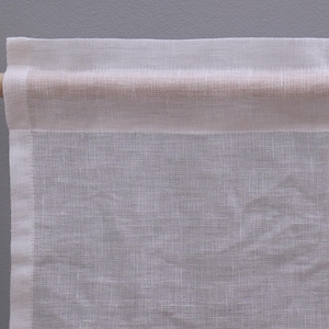 Peut inclure: Gros plan sur un panneau de rideau en lin blanc sur une tringle en bois. Le rideau a un design simple et minimaliste avec un tissage textur&eacute;. La tringle est de couleur marron clair et le fond est gris neutre.