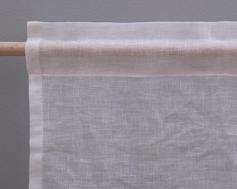 Peut inclure: Gros plan sur un panneau de rideau en lin blanc sur une tringle en bois. Le rideau a un design simple et minimaliste avec un tissage textur&eacute;. La tringle est de couleur marron clair et le fond est gris neutre.