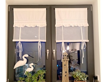 Puede incluir: Dos cortinas blancas de persiana romana con un ribete fruncido están colgadas en una ventana. La ventana está enmarcada en madera oscura. Las cortinas están subidas para revelar una vista de una habitación con una figurita de faro y plantas.