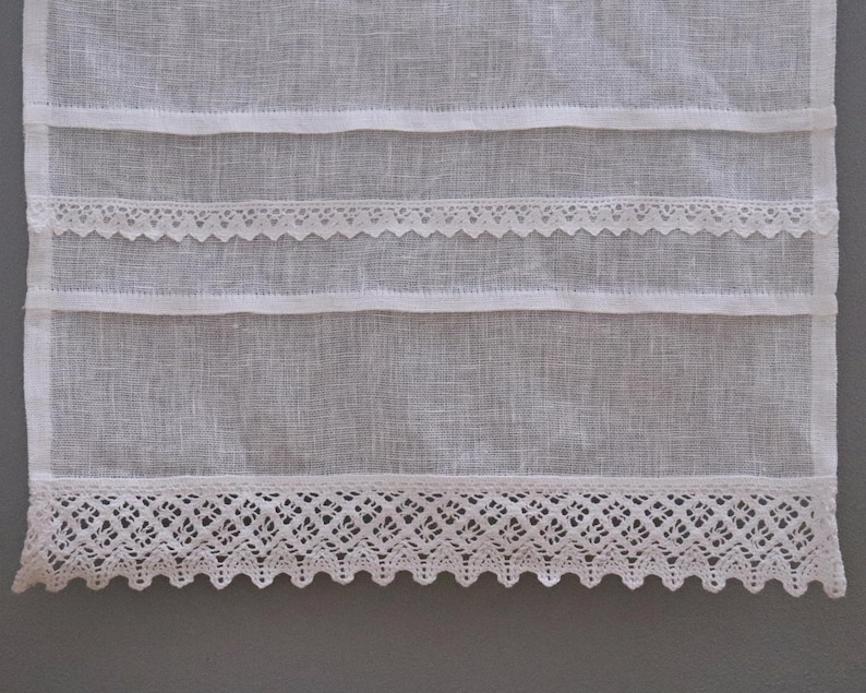 Peut inclure: Gros plan sur un textile en lin blanc avec des rang&eacute;es horizontales d'&eacute;l&eacute;ments d&eacute;coratifs. Le motif pr&eacute;sente des bandes de tissu et une d&eacute;licate garniture en dentelle. La dentelle a un bord festonn&eacute;, ajoutant une touche d'&eacute;l&eacute;gance.