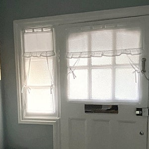 Può includere: Porta bianca con finestra e finestra più piccola a sinistra. Entrambe le finestre hanno tende bianche a pois con bordo a balze. La porta ha una fessura per la posta e una serratura. Una piccola mensola con una pianta è sopra la porta.