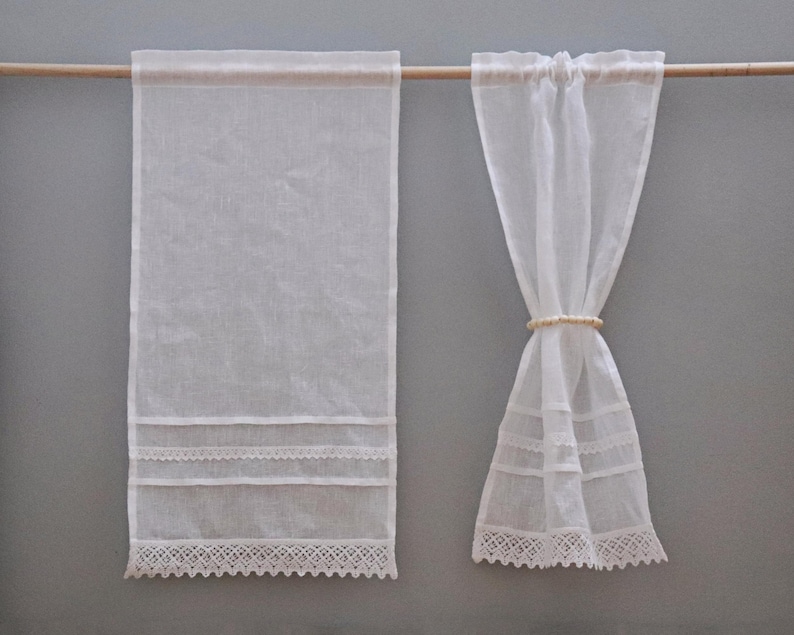 Peut inclure: Deux rideaux en lin blanc transparent suspendus &agrave; une tringle en bois. L'un est enti&egrave;rement d&eacute;ploy&eacute;, avec une bordure en dentelle d&eacute;taill&eacute;e en bas et des bandes de dentelle horizontales. L'autre est nou&eacute; avec un anneau clair.
