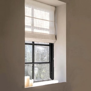 Può includere: Una tenda a pacchetto in lino beige è parzialmente sollevata su un telaio di finestra in metallo nero con più riquadri. La finestra rivela una vista di alberi all'esterno e la tenda presenta pieghe orizzontali che creano un aspetto stratificato.