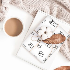May include: White wrapping paper with a repeating pattern of black gift boxes with bows. The paper is wrapped around a gift box and tied with twine. A cotton boll and a brown leaf are on top of the gift box.