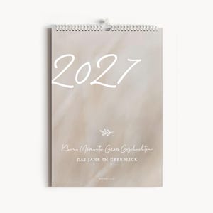 Könnte beinhalten: Ein Wandkalender für das Jahr 2027 mit beigefarbenem, weiß marmoriertem Hintergrund. Das Deckblatt zeigt den Text 2027 in weißer Schreibschrift, ein kleines Blattsymbol sowie den Text Kleine Momente Große Geschichten DAS JAHR IM ÜBERBLICK NORDEULE.