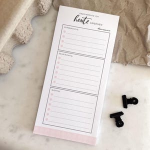 Könnte beinhalten: Ein weißer Notizblock mit rosa Akzenten und dem deutschen Text "Dass möchte ich heute angehen." Der Notizblock ist in drei Abschnitte unterteilt: "Super wichtig", "Sehr wichtig" und "Wichtig". Zwei schwarze Büroklammern liegen neben dem Notizblock.