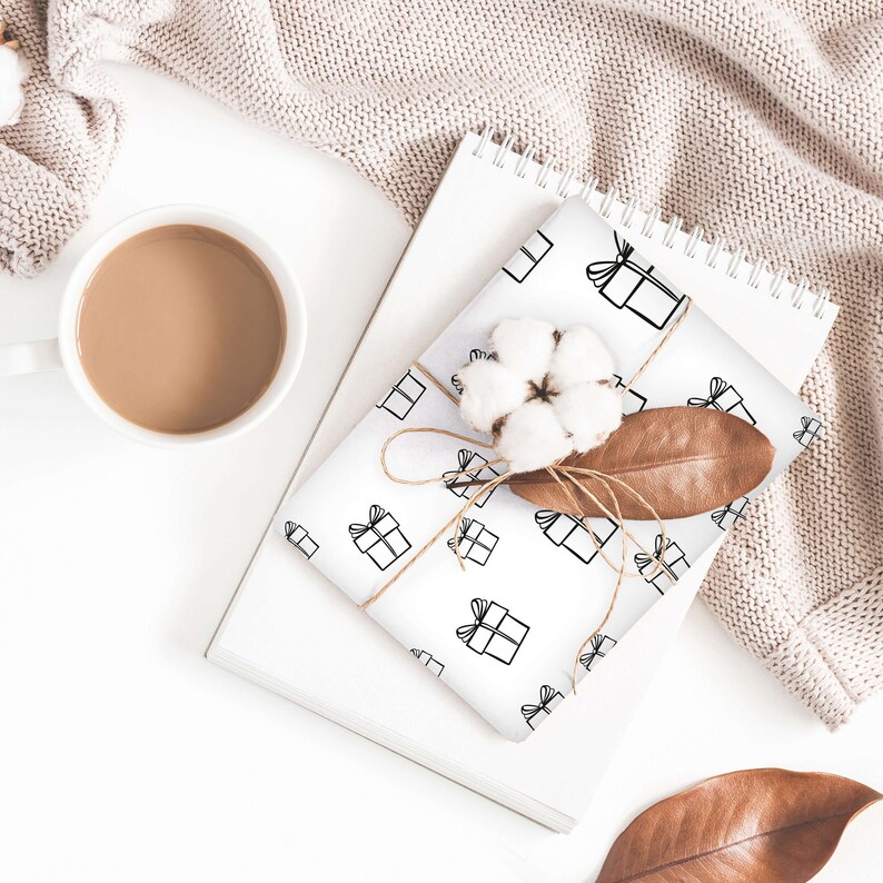 May include: White wrapping paper with a repeating pattern of black gift boxes with bows. The paper is wrapped around a gift box and tied with twine. A cotton boll and a brown leaf are on top of the gift box.