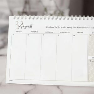 Könnte beinhalten: Ein weißer Tischkalender mit silberner Spiralbindung am oberen Rand. Die Seite zeigt den Monat August mit dem deutschen Text "Manchmal ist der größte Erfolg, den Schlüssel sofort zu finden" und ein Wochenraster von Montag bis Freitag.