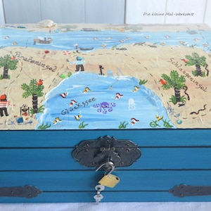 Könnte beinhalten: Eine blaue Holzschatztruhe mit einem bemalten Kartendesign auf dem Deckel. Die Karte zeigt eine Insel mit Palmen, einem See und dem Text "Glückesee". Die Truhe hat einen Metallverschluss und einen Schlüssel.