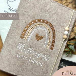 Könnte beinhalten: Ein beigefarbener Filzüberzug mit einem Regenbogenmuster und dem gestickten Text "Mutterpass Dein Name". Der Überzug hat eine weiße Schnur mit daran befestigten Holzperlen.