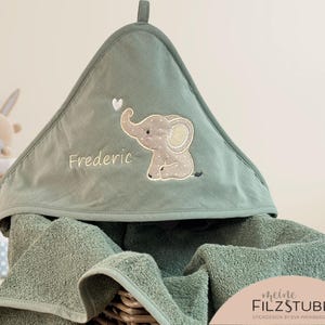 Könnte beinhalten: Ein grünes Kapuzenhandtuch mit einem gestickten Elefanten und dem Namen Frederic. Das Handtuch ist aus weichem, saugfähigem Material und perfekt für die Badezeit.