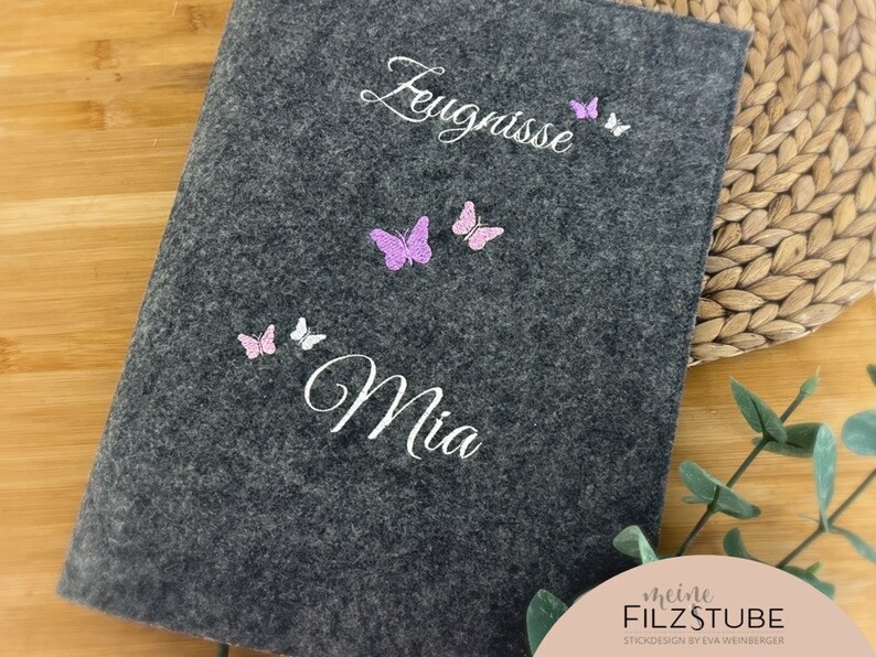 May include: Gray felt notebook with the text "Zeugnisse Mia" and pink and white butterfly embellishments. The notebook has a white label with the text "meine FILZSTUBE STICKDESIGN by EVA WEINBERGER".