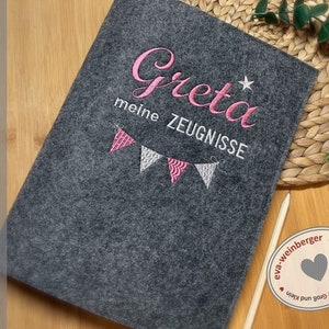 K&ouml;nnte beinhalten: Ein graues Filz-Notizbuch mit pinkfarbener Stickerei, auf dem "Greta meine Zeugnisse" mit einem Stern und einer Girlande aus rosa und wei&szlig;en Flaggen steht.