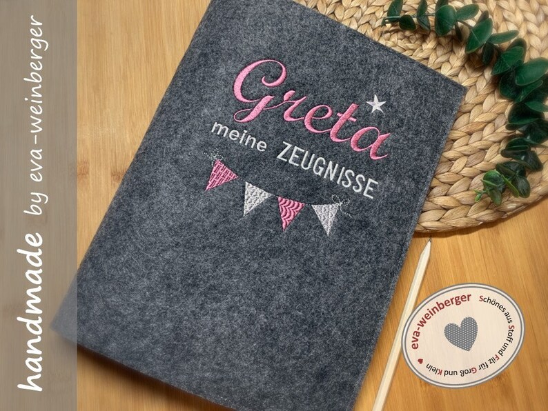 K&ouml;nnte beinhalten: Ein graues Filz-Notizbuch mit pinkfarbener Stickerei, auf dem "Greta meine Zeugnisse" mit einem Stern und einer Girlande aus rosa und wei&szlig;en Flaggen steht.