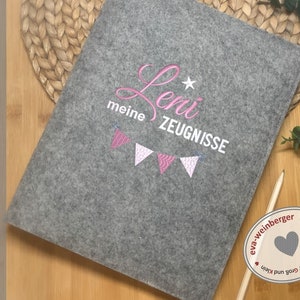 以下が含まれることがあります： グレーのフェルト製フォルダに、ピンクの刺繍で「Leni meine Zeugnisse」と星とピンクの旗の列が刺繍されています。
