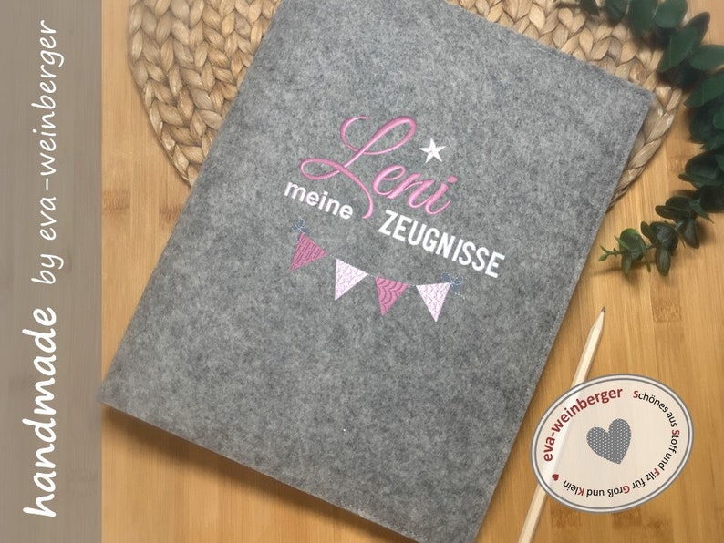 以下が含まれることがあります： グレーのフェルト製フォルダに、ピンクの刺繍で「Leni meine Zeugnisse」と星とピンクの旗の列が刺繍されています。