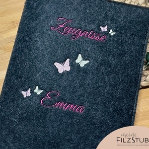 Puede incluir: Una carpeta de fieltro gris oscuro con mariposas bordadas en rosa y blanco y el texto "Leugnisse" y "Emma". La parte inferior de la carpeta tiene el texto "meine FILZSTUBE STICKDESIGN by EVA WEINBERGER".