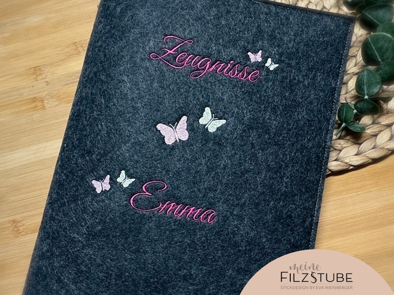 Puede incluir: Una carpeta de fieltro gris oscuro con mariposas bordadas en rosa y blanco y el texto "Leugnisse" y "Emma". La parte inferior de la carpeta tiene el texto "meine FILZSTUBE STICKDESIGN by EVA WEINBERGER".