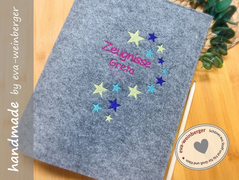 May include: A gray felt folder with a star design and the text "Zeugnisse Greta" embroidered in pink, blue, and yellow thread. The folder is handmade by eva-weinberger.