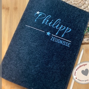 May include: A dark grey felt folder with the name "Philipp" embroidered in blue with two stars and the word "Zeugnisse" below. The folder is handmade by eva-weinberger.