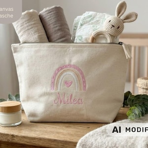 Puede incluir: Un neceser de lona beige con un arcoíris bordado en rosa y dorado, un corazón central y el nombre Milea en letra cursiva rosa. El neceser contiene dos paños de muselina doblados, un paño estampado y un pequeño juguete de conejo de ganchillo.