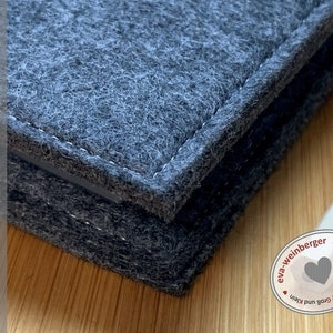 May include: Close-up of a grey felt notebook with a stitched edge. The notebook is on a wooden surface. A white pencil is visible in the background. The label on the notebook reads "handmade by eva-weinberger".