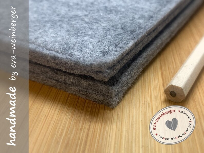 May include: Two layers of grey felt fabric with a stitched edge. The felt is folded in half and the edges are visible. The felt is on a wooden surface. The text "handmade by eva-weinberger" is visible on the left side of the image. The text "eva-weinberger Sch&ouml;nes aus Stoff und Filz f&uuml;r Gro&szlig; und Klein" is visible in a circle on the right side of the image.