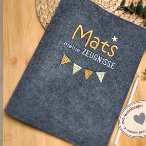 K&ouml;nnte beinhalten: Ein grauer Filzordner mit dem gestickten Text "Mats meine ZEUGNISSE" in Gelb und Wei&szlig;. Ein kleiner Stern ist &uuml;ber dem Namen gestickt. Der Ordner ist mit einer Girlande aus gelben und wei&szlig;en Flaggen verziert.