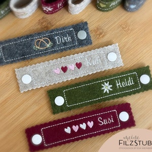 Puede incluir: Cuatro porta-nombres de fieltro con nombres y diseños bordados. Los nombres son Dirk, Kati, Heidi y Susi. Los diseños incluyen un pretzel, corazones, una flor y más corazones. Los porta-nombres son grises, beige, verdes y burdeos.