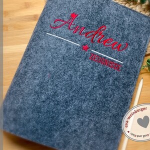 May include: Grey felt folder with red embroidered text "Andrew" and "Zeugnisse" with a red star above the name and below the text. The folder is handmade by eva-weinberger.