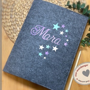 Könnte beinhalten: Grauer Filzordner mit dem Namen "Mara" in Lila mit einem Schwungmuster und pastellfarbenen Sternen bestickt. Der Ordner ist handgefertigt von eva-weinberger.