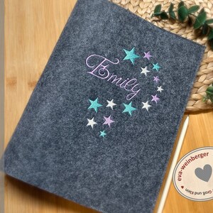 May include: Gray felt notebook with the name "Emily" embroidered in white thread with a design of teal and pink stars. The notebook is handmade by eva-weinberger.