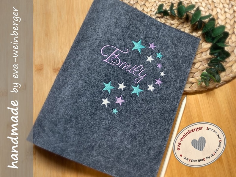 May include: Gray felt notebook with the name "Emily" embroidered in white thread with a design of teal and pink stars. The notebook is handmade by eva-weinberger.