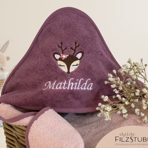Könnte beinhalten: Ein Kapuzenhandtuch in einem tiefen Lila mit einem rosa und weißen gestickten Reh mit einer Blumenkrone und dem Namen "Mathilda" darunter gestickt. Das Handtuch liegt in einem Weidenkorb mit einem rosa Handtuch darunter und einem Strauß getrockneter weißer Blumen.