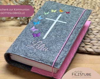 May include: Gray felt book cover with a pink border. The cover features a white cross with a circle of colorful butterflies around it. The name "Lilia" is embroidered on the cover. The text "Geschenk zur Kommunion GOTTESLOBHÜLLE" is visible at the top of the cover.