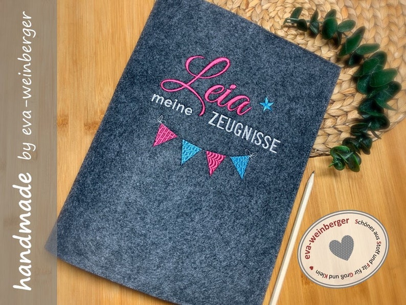 Pu&ograve; includere: Una cartella in feltro grigio con il nome "Leila" ricamato in rosa e bianco. La cartella ha anche una fila di bandierine colorate e il testo "meine Zeugnisse" ricamato in bianco.