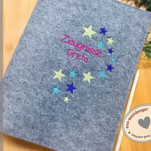 May include: A grey felt folder with a star design and the text "Zeugnisse Greta" embroidered in pink, blue, and yellow thread. The folder is handmade by eva-weinberger.