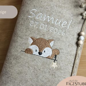 May include: Beige felt baby book cover embroidered with the name "Samuel" and the date "27.01.2026" in white. A brown and white fox is embroidered below the date, holding a star. The text "Filz: beige" is in the upper left corner.