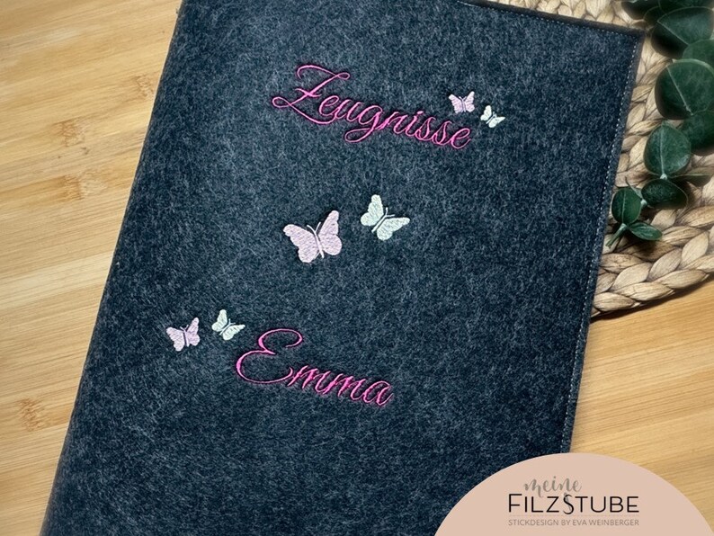 Puede incluir: Una carpeta de fieltro gris oscuro con mariposas bordadas en rosa y blanco y el texto "Leugnisse" y "Emma". La parte inferior de la carpeta tiene el texto "meine FILZSTUBE STICKDESIGN by EVA WEINBERGER".