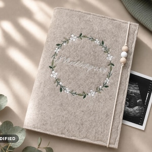 Puede incluir: Una funda de fieltro beige para cuaderno con una corona floral bordada en hilo verde y blanco. El centro contiene texto en cursiva. Una banda elástica vertical con tres cuentas de madera y blancas asegura la funda, sosteniendo una ecografía.