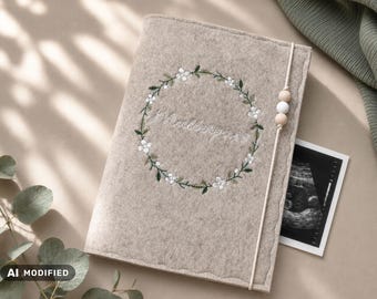 Puede incluir: Una funda de fieltro beige para cuaderno con una corona floral bordada que rodea el texto en cursiva "Madlepears". La funda se asegura con una banda elástica con dos cuentas de madera y una blanca, que sujeta una imagen de ecografía.