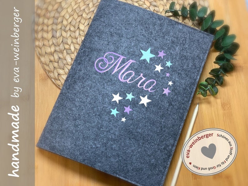 May include: Gray felt folder with the name "Mara" embroidered in purple with a swirl design and several pastel stars. The folder is handmade by eva-weinberger.