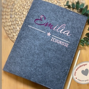 May include: Grey felt folder with the name "Emilia" embroidered in pink and a star. The folder has a white line and the text "Zeugnisse" embroidered in white below the name. The folder is handmade by eva-weinberger.