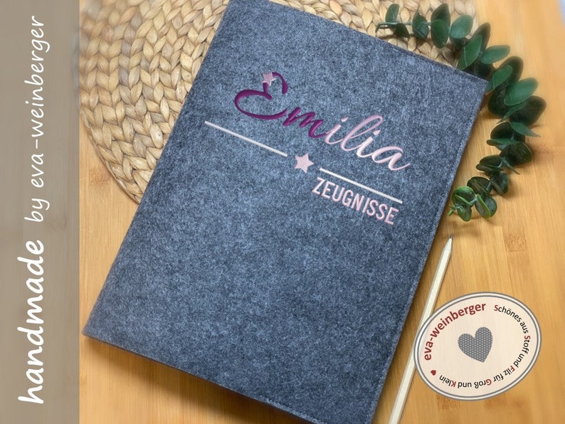 May include: Grey felt folder with the name "Emilia" embroidered in pink and a star. The folder has a white line and the text "Zeugnisse" embroidered in white below the name. The folder is handmade by eva-weinberger.
