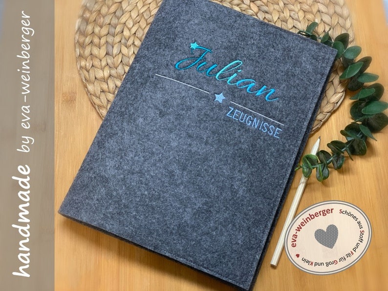 May include: Grey felt folder with the name "Julian" embroidered in blue and white with the word "Zeugnisse" below. The folder is on a wooden surface with a woven mat and a green plant.