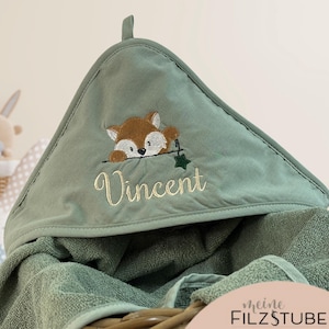 Puede incluir: Una toalla con capucha verde salvia para bebé con un diseño de zorro bordado y el nombre "Vincent". La toalla está doblada en una cesta, con un conejito de peluche y otras toallas en el fondo.