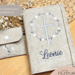 May include: A light beige felt book cover and small pouch set. The book cover features a cross within a floral wreath and the name "Leonie" with a date. The pouch has a floral embroidery and a button closure. The set is on a wooden surface.