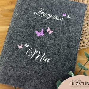 May include: Gray felt notebook with the text "Zeugnisse Mia" and pink and white butterfly embellishments. The notebook has a white label with the text "meine FILZSTUBE STICKDESIGN by EVA WEINBERGER".